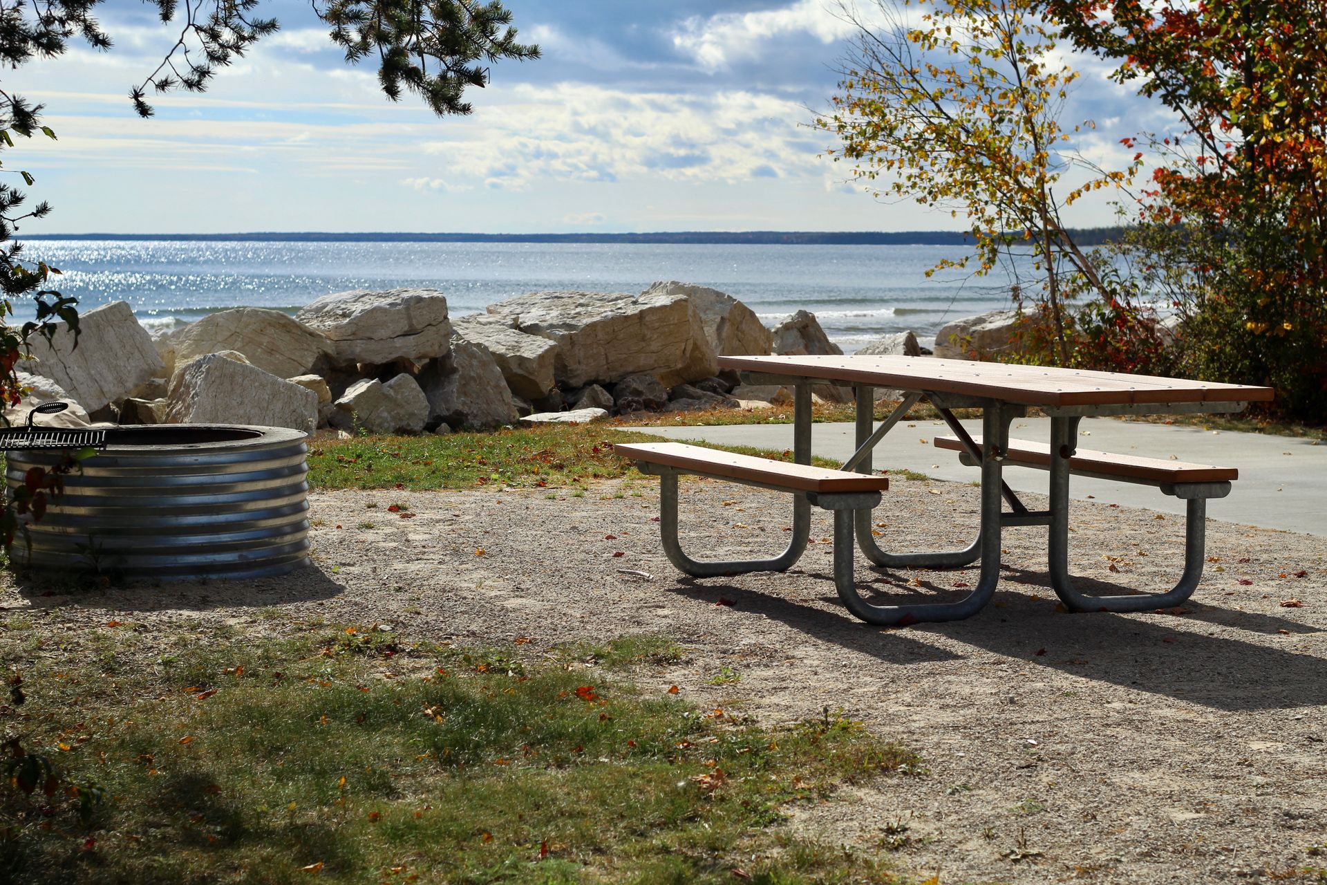 Manistique Lakeshore Campground: A serene outdoor scene featuring a picnic table by the beach, with rocks and gentle waves under a cloudy sky.