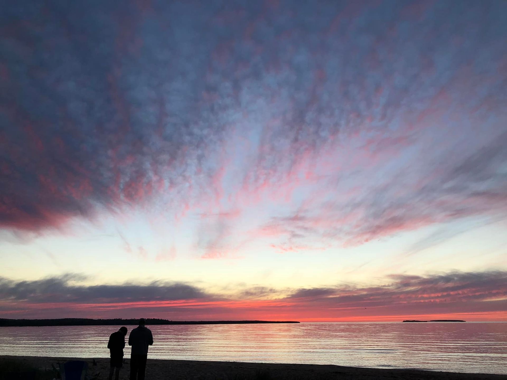 Munising Tourist Park Campground: A serene beach at sunset with vibrant pink and blue skies, silhouetted figures enjoying the view over calm waters.