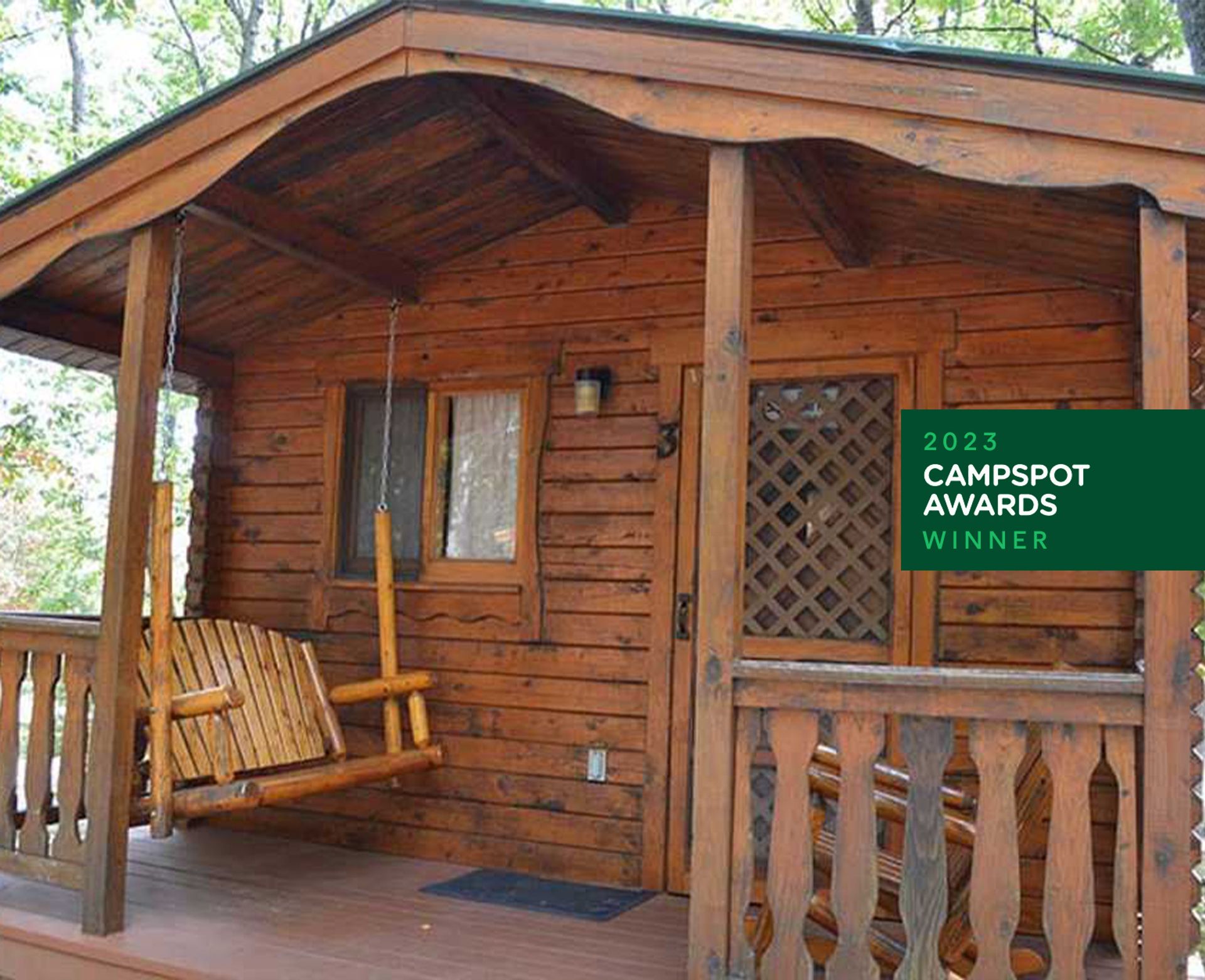 Indian River RV Resort, Indian River, Michigan: A cozy wooden cabin with a porch swing, showcasing rustic charm amidst nature, highlighted as a 2023 CampSpot Awards winner.